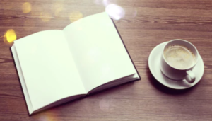 Open diary with blank pages sits on a wooden desk. A cup of coffee sits to the right. 