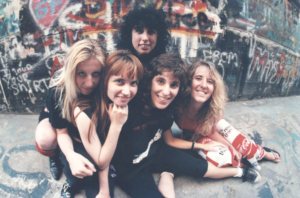 5 women posing on the ground, a wall of graffiti behind them.