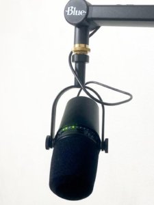 A close up of the Shure mic, pointing at the viewer, hanging from a mic arm. Green lights are displayed near the soft mic cover the user speaks into.